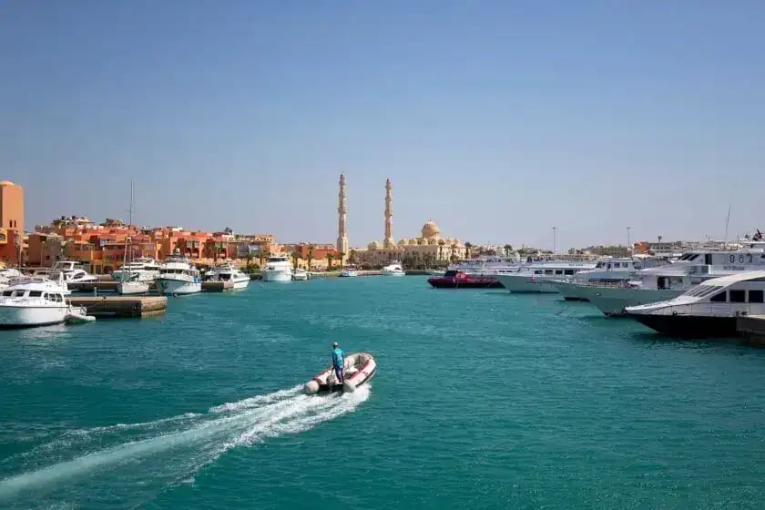 Speedboat entering Hurghada port, turquoise Red Sea channel flanked by moored yachts and mosque minarets