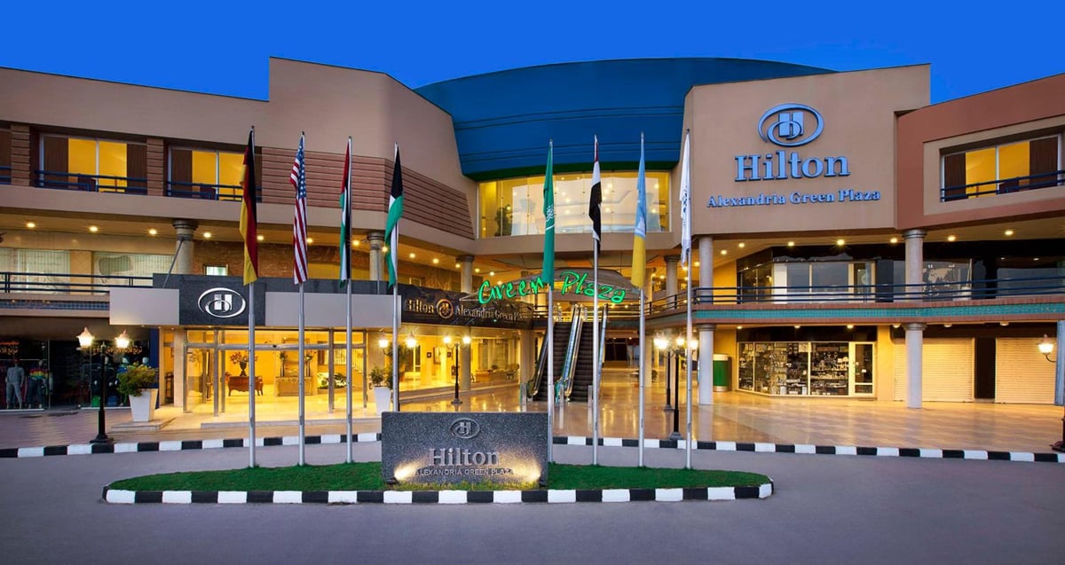 Hilton Alexandria Green Plaza hotel entrance with a glass canopy and flagpoles.