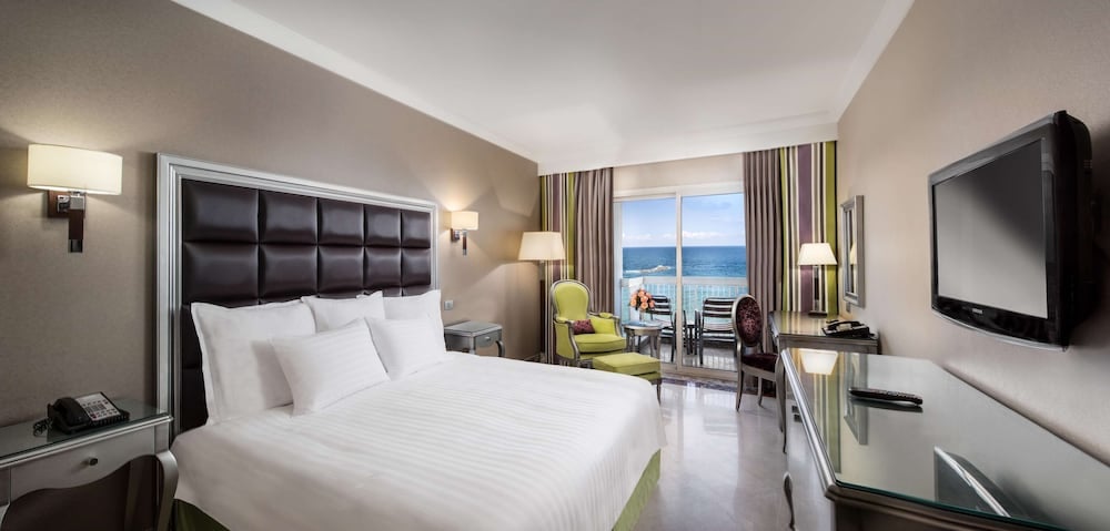 Hilton Alexandria Corniche guest room with a white bed and balcony overlooking the sea.