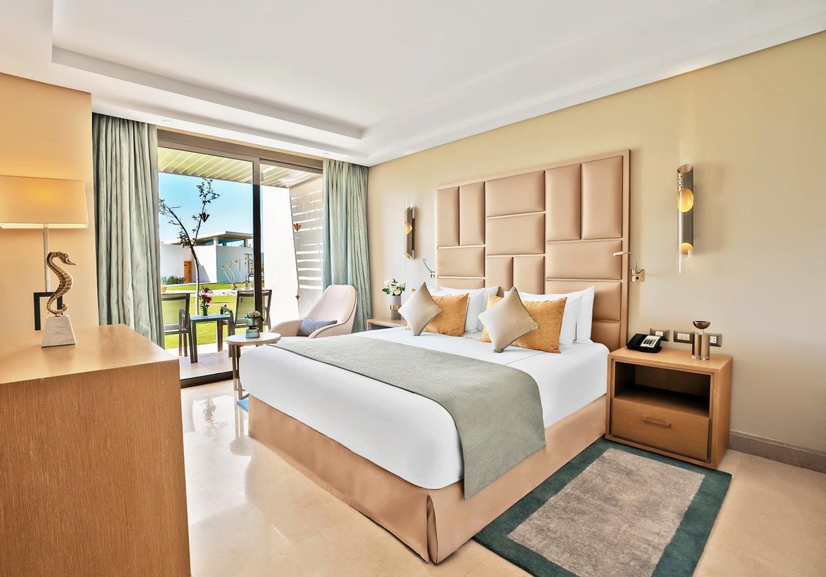 Rixos Premium Magawish Suites & Villas guest bedroom with an upholstered headboard and sliding balcony doors.
